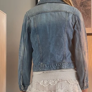 Ljusblå jeansjacka med knappar - Snygg ljusblå jeansjacka med klassisk krage och silverfärgade knappar framtill. Jackan har två bröstfickor med lock och långa ärmar med knapp vid mudden. Perfekt att slänga över en topp för en chill och trendig look.