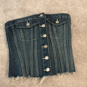 Jeanskorsett från Zara med knappar - Säljer en snygg mörkblå jeanskorsett från Zara i storlek XS. Toppen har silverfärgade knappar framtill, två fickor med lock och rå kant nedtill för en edgy vibe. Baksidan har smock för extra komfort och passform. Perfekt för dig som gillar en klassisk men unik jeans look på festen. 