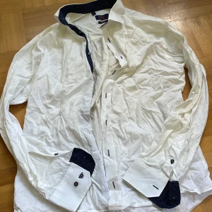 Vit slimfit skjorta från Oscar - Snygg vit skjorta från Oscar i slimfit-modell med mörkblå randiga detaljer på krage och manschetter. Skjortan har långa ärmar och knappar framtill. Perfekt för dig som gillar stilrena plagg med en twist. Storlek M (40).