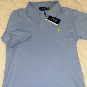 Ljusblå Polo Ralph Lauren t-shirt - Fräsch ljusblå t-shirt från Polo Ralph Lauren med klassisk krage och korta ärmar. T-shirten har den ikoniska gula broderade loggan på bröstet och två knappar vid halsen. Materialet är mjuk bomull som känns skönt mot huden.