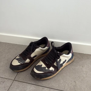 Valentino Rockrunner sneakers camouflage äkta! - Säljer ett par Valentino Rockrunner sneakers med svart och beige camouflagemönster, mörkblå och vinröda detaljer samt svarta snören. Skorna har platt sula och är tillverkade i mocka och skinn med coola nitar på hälen. Dom är köpta i valentino butiken men har tyvärr inte boxen kvar😔