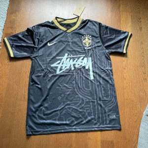 Svart Brasil x Stüssy x Nike fotbollströja - Limited edition svart Brasil fotbollströja från Nike och Stüssy med guldiga detaljer på krage och ärmslut. Broderat CBF-märke på bröstet, Stüssy-logga framtill och stor print på ryggen. Tillverkad i lätt, glansigt funktionsmaterial.