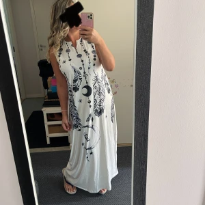 Vit långklänning med svart drömmarprint - Säljer en vit långklänning med coolt svart print av fjädrar, måne och drömfångare. Klänningen är ärmlös och har en avslappnad passform med liten krage och v-ringad hals. Perfekt för dig som gillar boho vibes och vill sticka ut med ett unikt mönster.Helt ny