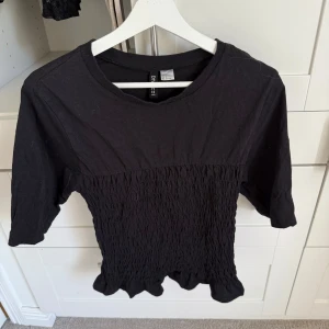 Svart smockad topp från Divided - Svart topp från Divided med smockad midja och volangkant nedtill. Modellen har rund hals och korta ärmar.