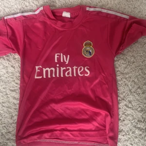 Rosa Real Madrid Benzema tröja - Snygg rosa Real Madrid fotbollströja med Benzema och nummer 9 på ryggen. Tröjan har korta ärmar, vita ränder på axlarna och klubbmärke broderat på bröstet. Materialet är lätt och andas, perfekt för träning eller match. Fly Emirates tryck på framsidan.