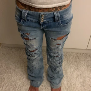 Ljusblå ripped jeans med bootcut - Säljer ett par ljusblå jeans med rejält slitna och trasiga detaljer framtill. Modellen har låg midja, fyra synliga knappar och klassiska fickor. Byxorna har bootcut-snitt och är tillverkade i jeansmaterial. Perfekt för dig som gillar en edgy och avslappnad stil. Midjemått: 41cm