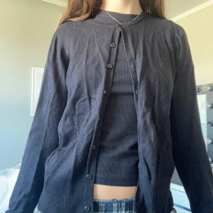 Svart kofta med knappar - En svart, klassisk kofta med rund hals och knappar hela vägen framtill. Koftan har långa ärmar och ribbade muddar vid ärmslut och nederkant. Materialet känns mjukt och lätt, perfekt att slänga över en topp. Enkel och stilren design som funkar till många olika outfits.