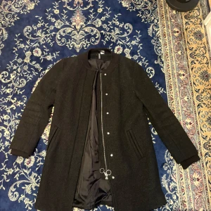 Svart rock med dragkedja och knappar - Svart rock från H&M med stilren design, rundad krage och långa ärmar. Jackan har både dragkedja med ringdetaljer och tryckknappar framtill samt två fickor på sidorna. Materialet känns mjukt och har en ullig yta som ger en schysst look.