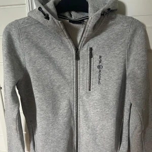 Grå hoodiejacka från Sail Racing - Säljer en grå hoodie från Sail Racing i storlek 160 med dragkedja framtill och på bröstfickan. Jackan har broderad logga på bröstet och huvan, samt justerbar snörning i huvan. Materialet är mjukt och känns som bomull med inslag av syntet vilket passar bra på chill dagar.