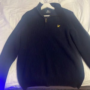 Svart stickad half zip från Lyle & Scott - Svart stickad tröja från Lyle & Scott med ribbad struktur och gul broderad logga på bröstet. Tröjan har half zip i halsen och långärmade ärmar. Perfekt för dig som gillar klassisk stil med en modern touch.