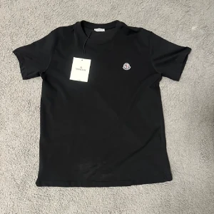 Svart Moncler t-shirt med logga - Svart t-shirt från Moncler med klassisk rund hals och korta ärmar. På bröstet sitter Monclers ikoniska logotyp i vitt och rött. T-shirten är gjord i mjuk bomull och har en clean, stilren look som passar till jeans eller shorts.