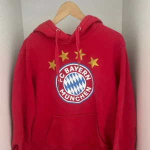 Röd FC Bayern München hoodie L - Säljer en röd hoodie från FC Bayern München i storlek L. Tröjan har klubbens stora logga framtill med fyra gula stjärnor ovanför, snörning vid huvan och en klassisk magficka. Perfekt för dig som älskar fotboll och vill visa ditt stöd för laget.
