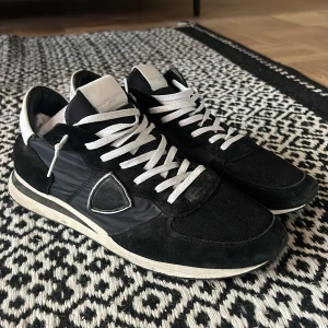 Svarta sneakers från Philippe Model - Snygga svarta sneakers från Philippe Model med vita detaljer och snörning. Skorna har en mix av mocka och syntet på ovansidan, svart/vit platt sula och rund tå. Ikonisk sköldlogga på sidan och vita skosnören ger en clean vibe. Perfekta när det blir lite kyligt ute. Hör av er om ni har några frågor.