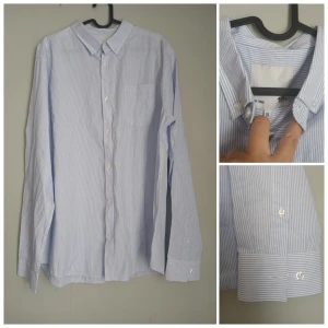 Klassisk Herrskjorta ljusblå/vit randig – stl XL - Klassisk långärmad herrskjorta i ljusblått och vitt med smala ränder. Tillverkad i ett lätt och bekvämt bomullsmaterial, perfekt för både vardag och arbete.  Storlek: XL  Färg: Ljusblå/vit randig  Modell: Button-down-krage, bröstficka, manschetter med knapp  Material: Bomull (skön och andas bra)  Skick: Mycket fint skick, sparsamt använd. Något skrynklig efter förvaring, men utan fläckar eller skador.