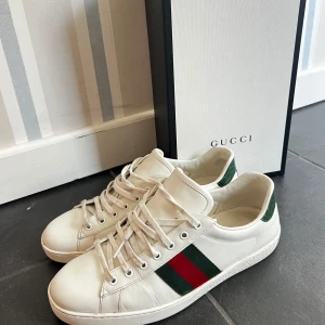 Gucci vita sneakers med gröna detaljer - Säljer ett par klassiska Gucci sneakers i vitt skinn med gröna och röda ränder på sidan samt grönt mönstrat skinn på hälen. Skorna har rund tå, platt sula och snörning framtill. Perfekt för dig som gillar lyxiga och stilrena sneakers med ikonisk design. En liten defekt på högra skon i fram, men syns knappt, se bild 3☺️innersula 29,5 cm 