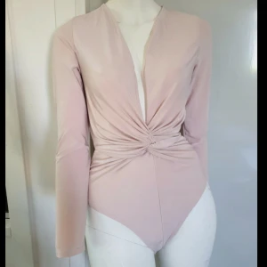 Ljusrosa omlott bodysuit med twist - Supersnygg ljusrosa bodysuit med omlott och twistad detalj i midjan. Djup v-ringning framtill och långa ärmar ger en clean och modern vibe. Stretchigt och mjukt material som sitter tight och smickrande. 