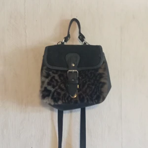 Svart ryggsäck med leopardpälsdetalj - Cool liten svart ryggsäck med leopardmönstrad päls på framsidan och snygga spänndetaljer i metall. Väskan har både handtag och axelremmar, samt ett lock med spänne och innerfack. Materialmix av skinn och päls ger en unik vibe.