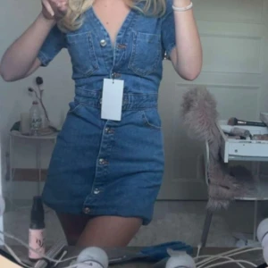 Zara  jeansklänning - Säljer en snygg blå jeansklänning med korta ärmar och v-ringad hals. Klänningen har framfickor på bröstet och knappar hela vägen framtill. Perfekt för dig som gillar denim och vill ha en cool och avslappnad stil.