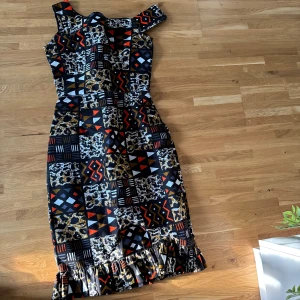 Mönstrad långklänning med volang - Snygg långklänning med coolt geometriskt och leopardinspirerat mönster i svart, vit, röd, beige och blå. Klänningen har en axelbandslös design och volangdetalj längst ner. Passformen är figurnära och materialet känns som bomull eller polyester.