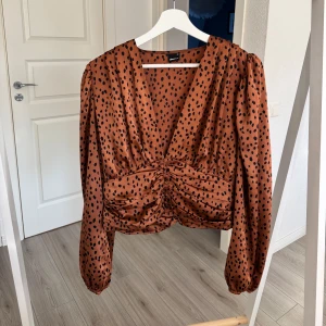 Brun prickig blus från Gina Tricot - Snygg brun blus med svarta ”fläckar” från Gina Tricot. Har djup v-ringning och rynkade detaljer framtill samt baksidan.Långa puffärmar och knappar vid framsidan. Materialet är glansigt och känns luftigt på.