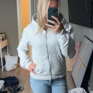 Ljusgrå hoodie med dragkedja - Säljer en ljusgrå hoodie med dragkedja framtill och huva. Tröjan har långa ärmar, ribbade muddar och två fickor fram. Perfekt för chill dagar eller när du vill ha något bekvämt och enkelt. Passar dig som gillar en clean och basic stil.