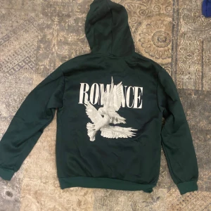 Mörkgrön hoodie med tryck Romance - Snygg mörkgrön hoodie med stor vit text 'ROMANCE' och två vita duvor tryckta på ryggen. Framtill finns samma text i mindre format. Tröjan har huva med snörning och en klassisk magficka. Perfekt för dig som gillar streetwear och coola prints.