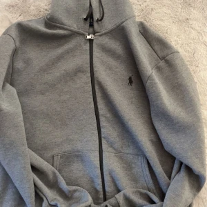 Grå hoodie från Polo Ralph Lauren - Säljer en grå hoodie från Polo Ralph Lauren med dragkedja och huva. Tröjan har svarta snören vid huvan, fickor framtill och den klassiska Polo-loggan broderad på bröstet. Perfekt för chill dagar och enkel att matcha med jeans väldigt bra skick. (PRISET KAN DISKUTERAS)