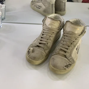 Saint Laurent höga sneakers i beige skinn - Unika höga sneakers från Saint Laurent i beige skinn med handskriven logga på sidan. Skorna har snörning, rund tå och platt sula. Tillverkade i Italien och har slitna detaljer som ger en cool vintagekänsla. Perfekt för dig som gillar statement pieces och streetstyle. Har box men ej nära för tillfället vid intresse kan jag fixa bilder.