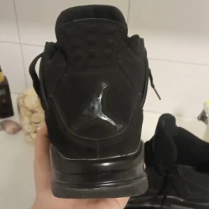 Svarta Nike Jordan 4 sneakers - Säljer ett par helsvarta Nike Jordan 4 sneakers med snygga meshpaneler och detaljer i plast. Skorna har snörning, platt sula och den ikoniska Jumpman-loggan på hälen. Ovandelen är i mocka och syntet, vilket ger en clean och modern vibe.