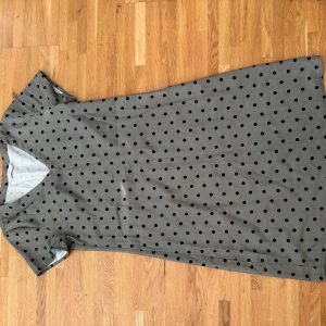 Grön prickig klänning med v-ringning - Snygg grön  klänning med svarta prickar och v-ringad hals. Modellen är rak och har korta ärmar, vilket ger en chill och enkel vibe. Perfekt för dig som gillar mönster och vill ha något som sticker ut lite. Materialet känns mjukt och lätt mot huden.