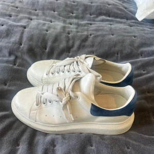 Alexander McQueen vita sneakers med blå mockadetalj - Säljer ett par Alexander McQueen sneakers i vitt med chunky platt sula och blå mockadetalj. Klassisk snörning, perfekt för dig som gillar stilrena och lyxiga sneakers. Logga på baksidan och så snygga för alla säsonger! 