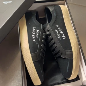 Svarta Saint Laurent sneakers med beige sula - Snygga svarta sneakers från Saint Laurent med beige platt sula och handskriven logga på sidan. Skorna har snörning, rund tå och är tillverkade i en mix av skinn och textil. Perfekt för dig som gillar stilrena och trendiga detaljer.