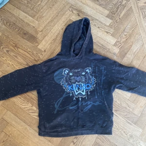 Svart hoodie med Kenzo tigertryck - Svart hoodie från Kenzo med blått tigertryck och Kenzo Paris-logga på bröstet. Tröjan har huva, lång ärm och ett diskret mönster med små vita streck över hela tyget. 