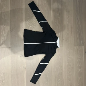 Svart långärmad konståknings tröja  - Svart långärmad tröja från ChloeNoel med vita ränder längs ärmarna och dragkedja framtill. Tröjan har en hög krage och är tillverkad i ett stretchigt syntetmaterial som passar perfekt för träning eller aktiva dagar. Snygg och sportig look med slim passform. Den saknar tre diamanter på den högra armen.💕