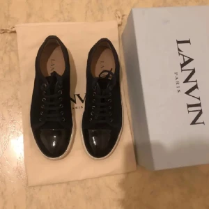 lanvins  - Snygga svarta sneakers från Lanvin med vit platt sula och lackad tå. Skorna har snörning och en stilren design med en mix av mocka och lackdetaljer. Perfekta för dig som gillar exklusiva och trendiga sneakers med en clean look.