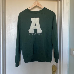 Grön sweatshirt med Athletics-tryck - Säljer en grön sweatshirt med stort vitt A och Athletics-tryck på bröstet. Tröjan har rund hals, långa ärmar och ribbade muddar. Perfekt för dig som gillar sportig och chill stil. Materialet känns som mjuk bomull och passar sköna dagar.