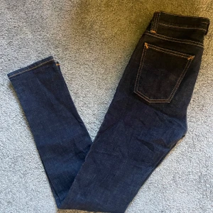 Mörkblå skinny jeans från Nudie Jeans - Snygga mörkblå skinny jeans från Nudie Jeans i ekologisk bomull. Klassisk femficksmodell med orange kontrastsömmar och läderpatch med logga bak. Passar dig som gillar stilrena och tajta jeans med modern vibe.