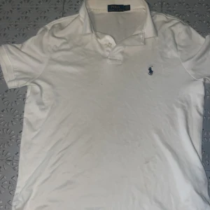 Vit Polo Ralph Lauren piké - Vit piké från Polo Ralph Lauren med klassisk pikékrage och korta ärmar. Diskret blå logga broderad på bröstet. Tillverkad i mjuk bomull som känns skön mot huden. Perfekt för dig som gillar stilrena och tidlösa plagg. Passar S