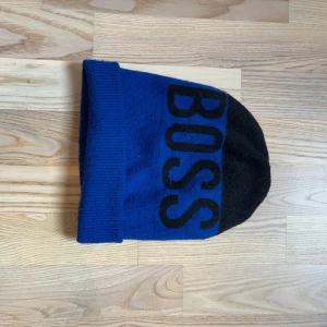 Blå och svart mössa från BOSS - Snygg mössa från BOSS i blått och svart med stor BOSS-logga framtill. Klassisk form med uppvikt kant och mjukt stickat material som sitter skönt på huvudet. Perfekt för dig som vill ha en stilren accessoar med tydlig branding.
