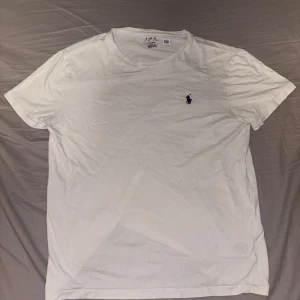 Vit t-shirt från Polo Ralph Lauren - Vit t-shirt från Polo Ralph Lauren med klassisk rund hals och blå broderad logga på bröstet. Tillverkad i mjuk bomull, perfekt för en clean och enkel stil. Passformen är normal och ärmarna är korta. Enkelt plagg som funkar till jeans eller shorts.