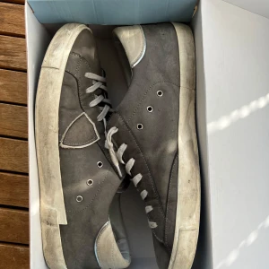  Philippe Model Skor - Säljer ett par grå Philippe Model sneakers med vit sula och ljusgrå snörning. Skorna har en rund tå, platt sula och detaljer i mocka. Klassisk design med logotyp på sidan och insida i ljusgrått. Perfekt för dig som gillar stilrena och snygga sneakers.