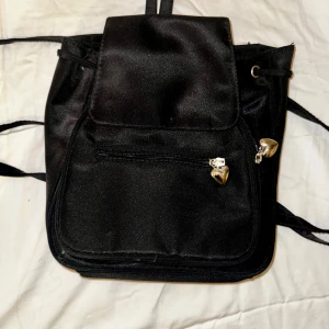 Svart satin ryggsäck med hjärtdetaljer - Snygg svart ryggsäck i satin med glansig finish och silverfärgade hjärtformade dragkedjor. Väskan har snörning upptill, flera fack med dragkedja och justerbara axelremmar. Perfekt för dig som vill ha en unik och stilren väska med lite extra detaljer.