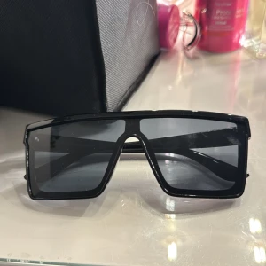 Svarta rektangulära solglasögon - Säljer ett par svarta solglasögon med breda rektangulära bågar och mörka linser. Glasögonen har en modern och edgy look som passar dig som vill sticka ut. Perfekta för att ge din outfit en cool vibe.