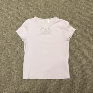 Ljuslila ribbad topp med knyt från Lindex - Supersöt ljuslila ribbad topp från Lindex med korta ärmar och en liten knyt-detalj vid halsringningen. Tillverkad i mjuk ekologisk bomull som känns skön mot huden. Perfekt för dig som gillar enkel men gullig stil.