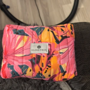 Glossybox blommig quiltad handväska - Säljer en färgstark och quiltad handväska från Glossybox med blommigt mönster i rosa, orange, gul och mörkblå toner. Väskan har en praktisk dragkedja upptill och är tillverkad i mjuk bomull. Perfekt för att hålla ordning på dina småsaker.