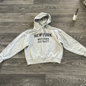 Grå hoodie med New York-tryck 157 XS - Grå hoodie från 157 i storlek XS med svart texttryck 'NEW YORK MIDTOWN DISTRICT' på bröstet. Tröjan har huva med snörning och ribbade muddar vid ärmslut och nederkant. Perfekt för dig som gillar streetstyle och chill vibes.