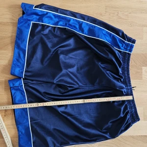 Basketshorts medium/large - Snygga marinblå sportshorts med breda blå paneler och vita kantlinjer längs sidorna. Shortsen har elastisk midja med snörning för skön passform och är gjorda i glansigt syntetmaterial som känns lätt och luftigt. Perfekta för träning eller chill.