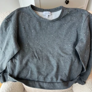 Grå sweatshirt med stenar & Other Stories - Mysig grå sweatshirt från & Other Stories Paris Atelier, dekorerad med små glitterstenar på framsidan. Tröjan har rund hals och långa ärmar, insidan är mjukt fodrad för extra komfort. Perfekt för chill dagar och när du vill ha en enkel men snygg look.