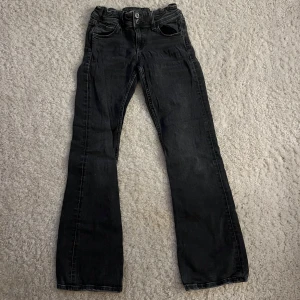 Svarta bootcut jeans med fickdetaljer - Snygga svarta jeans med bootcut passform och klassiska fem fickor. Baktill finns fickor med lock och knappdetaljer som ger extra stil. Jeansen har normal midja och är tillverkade i mjukt denimtyg som sitter skönt hela dagen. Storlek 152 cm 