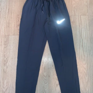 Mörkgråa dri fit byxor från Nike M - Svarta träningsbyxor från Nike i storlek M med klassisk vit swoosh-logga på benet. Byxorna har elastisk midja med snörning för skön passform och är gjorda i lätt polyester som andas. Perfekta för gymmet eller chill hemma.